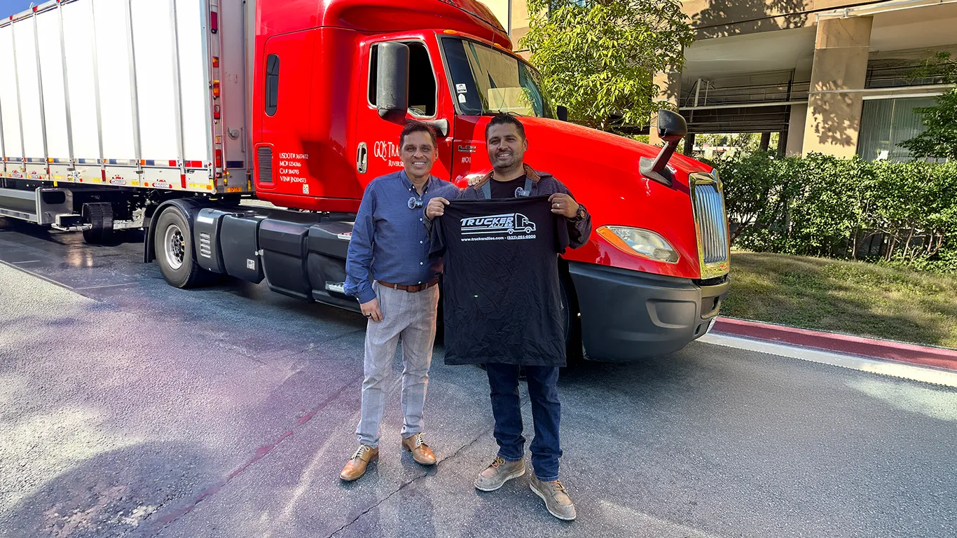 Jorge H., cliente de Trucker Allies, junto a un miembro del equipo de Allies frente a un camión rojo de GQ’s Transport, sosteniendo en alto una camiseta de Trucker Allies.
