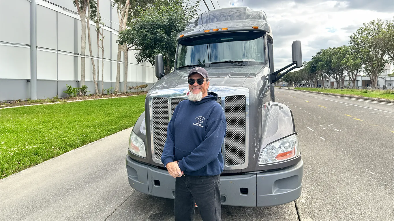 Larry H., cliente de Trucker Allies, con lentes de sol y sudadera azul marino, frente a la parrilla cromada de un camión gris.
